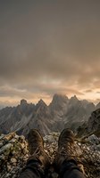 Hiker boots resting on rocky mountain ledge with dramatic alpine peaks and moody sunset sky creating adventurous outdoor exploration atmosphere.