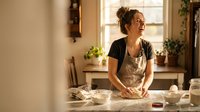 Young woman baker kneading dough in warm sunlit kitchen wearing apron, smiling while preparing homemade bread for baking and cooking activities.