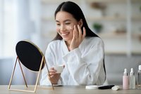 Young Asian woman applying skincare cream while looking in mirror during morning beauty routine at home vanity table with cosmetic products.
