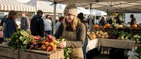 Young blonde woman in beige knit hat and apron selecting fresh organic vegetables at outdoor farmers market. Local food shopping and sustainable living.