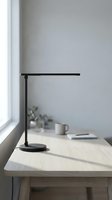 Modern black desk lamp on light wood table by window with minimalist workspace setup and natural daylight for office or home study environment.
