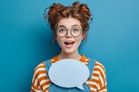Surprised young woman with curly red hair and round glasses holding blank speech bubble against blue background for social media or communication concept.