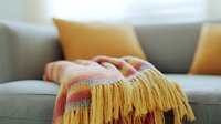 Cozy knitted throw blanket with yellow fringe on gray sofa, warm orange and pink stripes pattern, decorative cushions in background, home interior decor.
