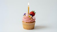 Vanilla cupcake with pink frosting, lit birthday candle, fresh strawberry, purple flower decoration and yellow sprinkles on white background for celebrations.