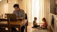 Mixed-race father working on laptop at home office desk while two young children play quietly on floor nearby in warm, sunlit living room.
