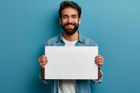 Smiling bearded man in casual denim shirt holding blank white poster against blue wall background. Great for advertising, announcement or promotion mockup.