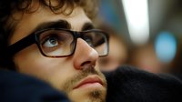 Young bearded man wearing glasses gazes upward thoughtfully in dramatic lighting, close-up portrait showing contemplative expression and modern eyewear.