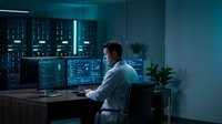 Young Caucasian male IT specialist working late at night in modern data center with multiple monitors displaying code and system analytics in blue ambient lighting.