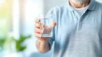 Senior Caucasian man in light blue polo shirt holding clear glass of pure water against blurred background, promoting healthy hydration and wellness.