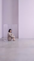 Young Asian woman sitting inside a transparent acrylic box in a minimalist lavender studio, expressing loneliness, isolation, and mental health concepts.