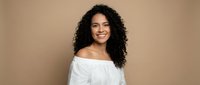 Young mixed-race woman with curly hair smiling warmly in white off-shoulder top against neutral beige background for lifestyle and portrait photography.