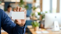 Businessman in dark blue shirt holding blank white business card toward camera in modern office environment with blurred colleagues and plants in background.