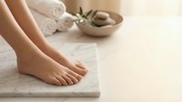 Soft female feet on marble surface with spa towels and stones in background, evoking relaxation and skincare wellness.