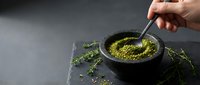Hand stirring vibrant green matcha powder in black bowl with wooden spoon on dark slate surface surrounded by fresh tea leaves for culinary preparation.