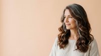 Young woman with long wavy dark brown hair and fair complexion wearing light gray casual top, smiling while looking to the side against warm beige background for portrait and lifestyle use.