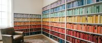 Modern home library with color-coordinated books arranged by rainbow spectrum on floor-to-ceiling shelves, leather armchair and natural window light.