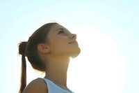 Young Asian woman with eyes closed facing sunlight in peaceful meditation pose, wearing light blue tank top against bright sky background during golden hour.