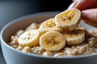 Fresh banana slices being added to creamy oatmeal porridge in white ceramic bowl, close-up view showing texture and natural breakfast preparation.