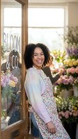 Young woman with curly dark hair and warm smile standing in flower shop doorway wearing floral dress and lavender cardigan, surrounded by fresh blooms and greenery for small business, retail, or florist concepts.