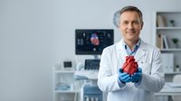 Middle-aged Caucasian male cardiologist in white coat holding anatomical heart model in modern medical office with cardiac monitor displaying heart rhythm.