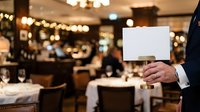 Businessman holding blank white card in upscale restaurant with elegant dining setup and warm ambient lighting, suitable for hospitality and business marketing.