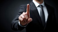 Businessman in formal suit pointing finger upward against dark background, demonstrating leadership and decision-making in corporate environment.