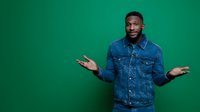 Young African American man in denim jacket shrugging with open palms against vibrant green background, expressing confusion or uncertainty.