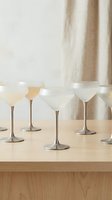 Elegant frosted glass cocktail glasses with metallic stems arranged on wooden surface in minimalist neutral interior setting for entertaining.