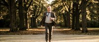 Mature male professional in business attire jogging through tree-lined park path during golden hour, promoting fitness, wellness, and active lifestyle for corporate health campaigns.