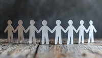 Paper cutout figures holding hands in row on wooden surface, symbolizing unity, teamwork and community support. Gradient light effect adds depth to composition.