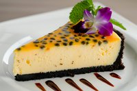 Passion fruit cheesecake slice with chocolate cookie crust, garnished with fresh orchid and mint leaf, served on white plate with chocolate sauce drizzle.