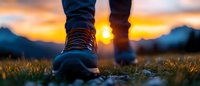 Hiking boots on grass at sunset with dramatic orange sky and mountain silhouettes in background, close-up panoramic view capturing outdoor adventure moment.