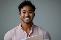 Young Asian male professional with bright smile wearing pink button-down shirt holding documents against neutral gray background, representing success and confidence.