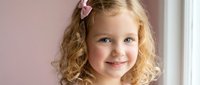 Young blonde Caucasian girl with curly hair and pink bow smiling at camera in bright indoor setting for children photography and family portraits.