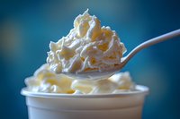 Creamy whipped topping on silver spoon above white container, macro shot with soft blue background showing texture and peaks of fresh dairy dessert.