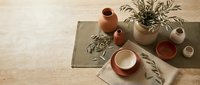 Minimalist ceramic tableware set with terracotta bowls and neutral vases arranged on natural linen placemats with dried botanical elements on wooden surface.