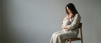 Young Caucasian mother in linen dress sits on chair, tenderly holding sleeping newborn against chest, soft gray studio background.