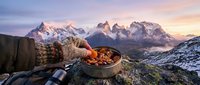Hiker holding bowl of hot stew with snow-covered mountain peaks at sunrise, outdoor camping meal in wilderness alpine landscape.