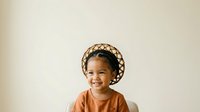 Young child with medium brown skin tone and dark curly hair wearing an orange shirt and patterned beige bonnet, smiling at camera against neutral background for portrait and lifestyle use.