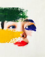 Asian woman's face partially revealed through vibrant brush strokes of green, yellow, blue and red paint, creating an artistic portrait on white background.