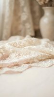 Delicate cream lace fabric with ornate scrollwork pattern draped softly, suitable for wedding gowns, bridal wear, formal occasions, and luxury textile design projects.
