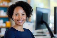 Young African American businesswoman with natural curly hair smiling confidently in modern office environment, wearing navy blue top, professional workplace portrait.