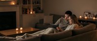Couple relaxing together on couch reading tablet in cozy living room with warm candlelight and fireplace creating intimate evening atmosphere at home.
