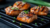 Grilled salmon fillets glazed with teriyaki sauce on black grill grates, garnished with sesame seeds and fresh chopped green onions, close up view of juicy fish.