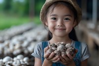 Young Asian girl in denim overalls and straw hat holding fresh mushrooms, smiling warmly at camera. Rural farm setting with natural bokeh background.