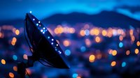 Satellite dish silhouette against blurred city lights at dusk creates dreamy bokeh effect with orange and blue glowing dots in twilight urban landscape.