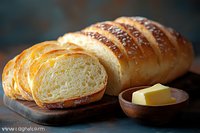 Fresh baked artisan bread loaf sliced on wooden board with butter in rustic bowl against dark background, warm natural lighting highlights texture.