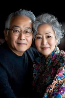 Senior Asian couple portrait showing warmth and connection. Elderly woman in colorful floral dress and man in dark sweater with glasses share genuine smiles against dark background.