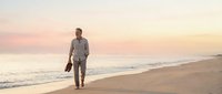 Mature man in casual business attire walking on sandy beach at sunset, holding shoes, contemplating career change or retirement lifestyle transition.