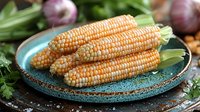 Fresh raw corn cobs with golden kernels arranged on rustic turquoise ceramic plate, surrounded by garlic and herbs, moody food photography setup.